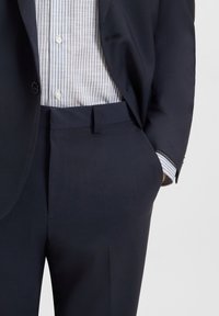Marineblauwe colbert met één knoop, gecombineerd met een lichtblauw en wit gestreept overhemd. De broek heeft een rechte voorkant en zijzakken.