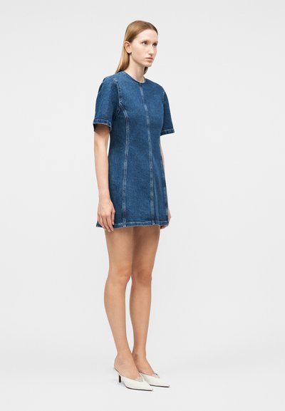 Γυναίκα που στέκεται φορώντας ένα κοντομάνικο, μπλε τζιν φόρεμα πάνω από το γόνατο και λευκά παπούτσια με μυτερή μυτούλα και χαμηλό τακούνι, σε απλό φόντο.