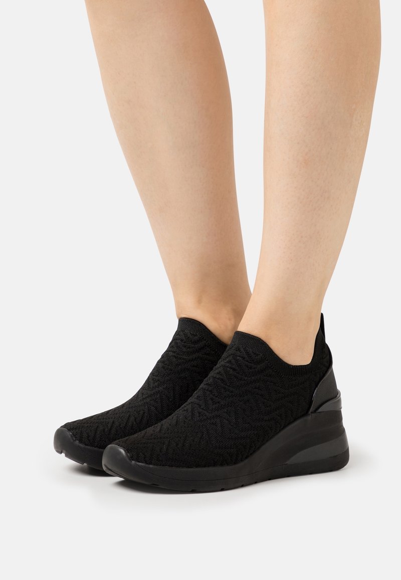 Baskets noires sans lacets avec tiges en maille texturée et semelles compensées portées à pieds nus sur un fond blanc.
