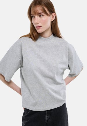 Jeune femme aux cheveux bruns raides, portant un t-shirt gris clair à manches courtes et col montant, ainsi qu'un pantalon noir, regardant vers sa gauche.