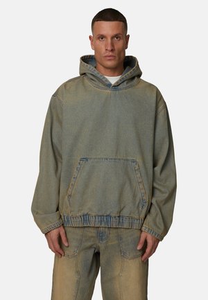 Uomo che indossa una felpa con cappuccio in denim verde-blu sbiadito e jeans abbinati, in piedi davanti a uno sfondo chiaro e uniforme, rivolto verso la fotocamera.