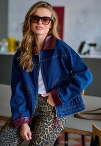 Džínová bunda s hnědým koženým límcem, zapínáním na zip a kontrastními bordó manžetami, nošená s kalhotami s leopardím vzorem.