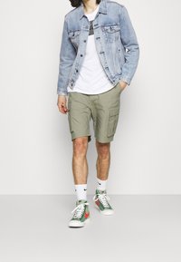 Ljusblå denimjacka över en vit t-shirt, gröna cargoshorts, vita och gröna sneakers samt vita strumpor med en svart logotyp.