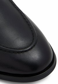 Chaussure en cuir noir avec une texture lisse, bout arrondi et des coutures visibles autour du bord. Semelle basse.