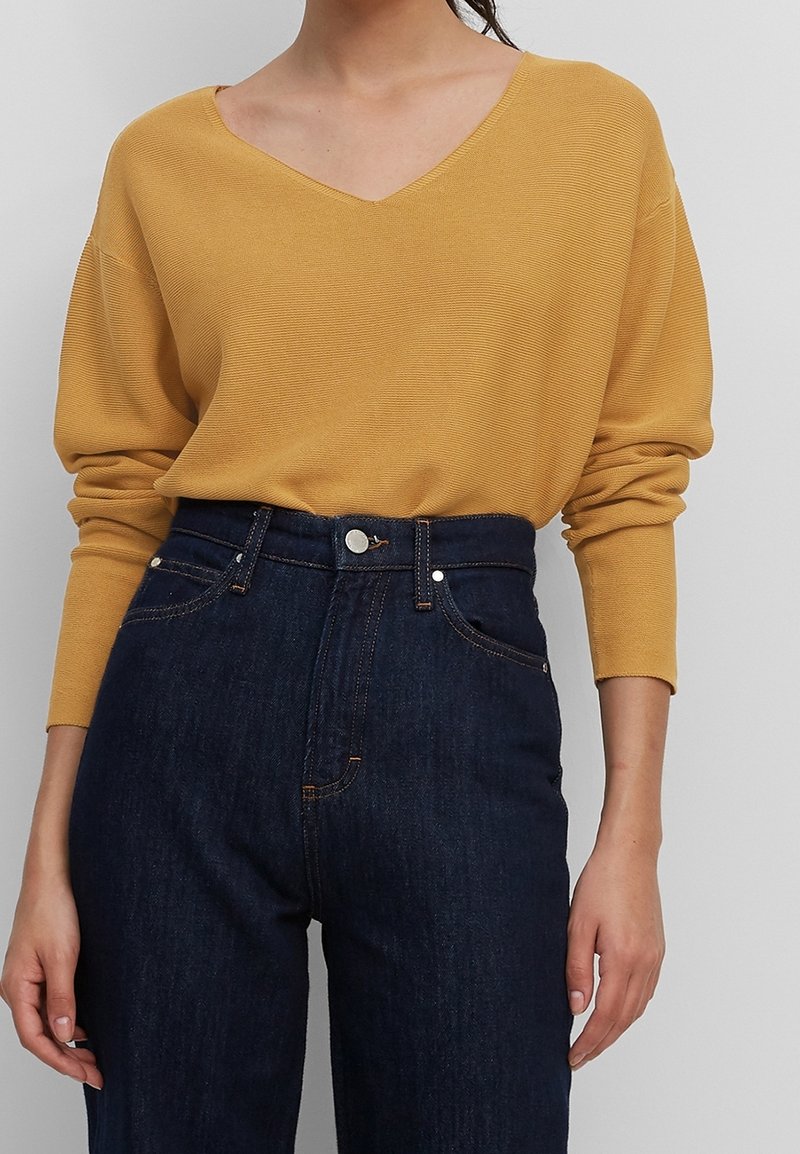 Maglione giallo senape a scollo a V con maniche lunghe, texture a coste, abbinato a jeans di denim blu scuro a vita alta con chiusura a bottone.