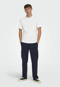 Weißes Crew-Neck T-Shirt, marineblaue Cargo-Hosen mit Seitentaschen, schwarze Sneaker mit weißen Sohlen. Einfaches, lässiges Outfit in einer Studio-Umgebung.