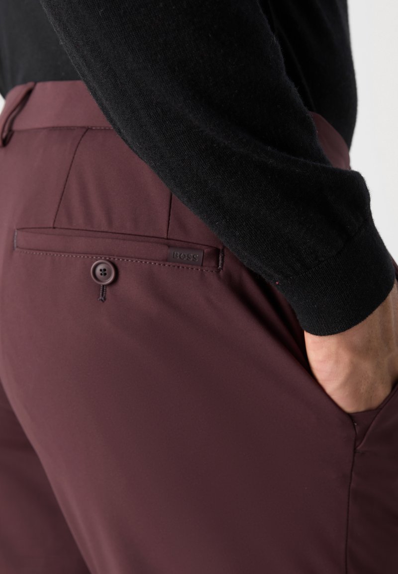 Burgunderfarbene Hosen aus glattem Stoff mit einer knöpfbaren Gesäßtasche; getragen mit einem schwarzen strukturierten Pullover. Besitzt ein dezentes Logoschild.