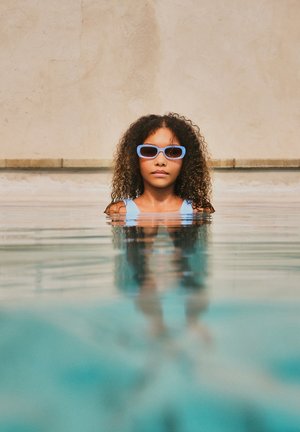 Γυναίκα με σγουρά μαλλιά, που φοράει γαλάζια γυαλιά ηλίου και μαγιό, στέκεται μέχρι το στήθος σε μια πισίνα, με ουδέτερο τοίχο στο φόντο.
