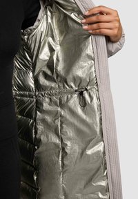 Gaiši pelēks mētelis ar spīguļojošu metāla sudraba oderi, ar quilted tekstūru un regulējamu aukliņu jostas vietā.