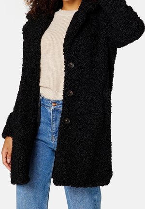 Manteau teddy noir texturé avec un col en V, des boutons et des manches longues, porté sur un pull en tricot crème et un jean bleu.