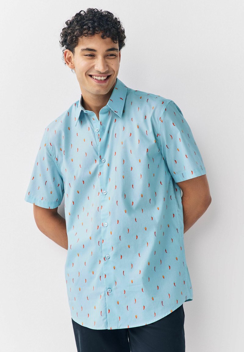 Ung mand smilende, iført en lyseblå kortærmet skjorte med knapper og et rødt chili pebermønster, stående mod en hvid baggrund.