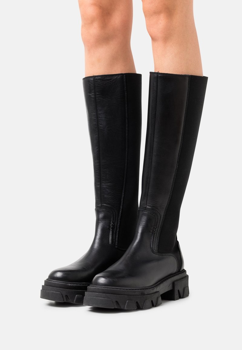 Bottes en cuir noires montant jusqu'au genou avec semelles crantées épaisses, portées par une personne debout devant un fond neutre.