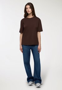 Brązowa bawełniana koszulka z okrągłym dekoltem i krótkimi rękawami, w zestawieniu z niebieskimi dżinsami. Koszulka ma luźny krój i prosty design.