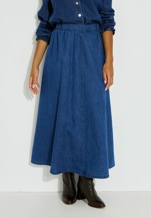 Femme portant une longue jupe en jean bleue, une chemise bleu foncé boutonnée avec des manches froncées, et des bottines noires.