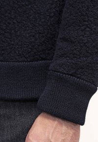 Marinblå fleece-tröja med manschetter av texturerat, fuzzy material. Har en mörk, ribbad nederkant, vilket visar en smidig övergång mellan material.