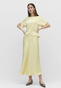 Satin to-delt outfit i lys gul; kortærmet top med løs pasform, lang nederdel med blød drapering, parret med mønstrede sko.