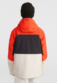 Chaqueta con bloques de color en naranja brillante, azul marino y crema. Presenta una capucha, corte relajado y material liso e impermeable.