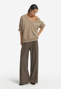 Przesadny beżowy sweter z dekoltem w serek i opadającymi ramionami, zestawiony z szerokimi brązowymi spodniami w kratę. Gładka tekstura i luźny krój.