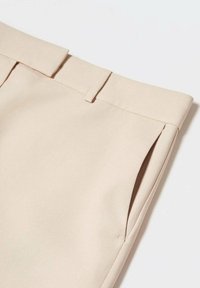 Pantalon beige ajusté avec passants pour ceinture et poches latérales, montrant la texture du tissu et des détails de couture soignés.