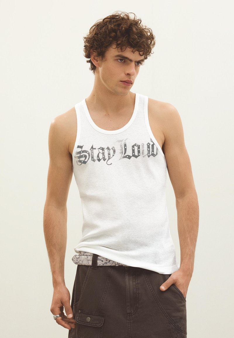 Jeune homme aux cheveux bouclés portant un débardeur blanc imprimé "Stay Loud" et un pantalon foncé, une main dans la poche.