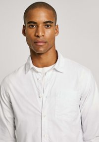 Camicia bianca con bottoni, dal tessuto morbido, dotata di taschino sul petto e colletto classico. Design pulito e minimale su uno sfondo neutro.
