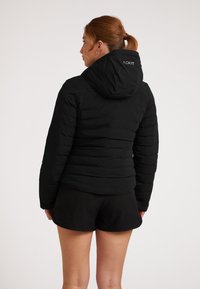 Sort pufferjakke med hætte, quiltet design og broderet logo på ryggen. Båret med sorte shorts. Glat, mat tekstur.