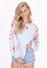 Maglietta azzurra chiara con maniche a fiori a motivi in rosa e arancione; texture a coste, scollatura rotonda e vestibilità rilassata. Abbinata a pantaloni shorts in denim strappato.