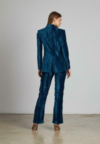 Il completo di velluto turchese presenta un blazer sartoriale con linee testurizzate e pantaloni dritti abbinati. La vista posteriore mostra una vestibilità snella e una struttura elegante.