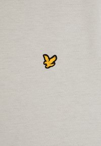 Light gray t-shirt with a textured, embroidered black and yellow bird logo on the left chest area. Soft cotton blend material.