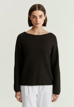 Femme aux cheveux courts et bruns portant un pull en maille noir et un pantalon blanc, debout devant un fond clair uni.