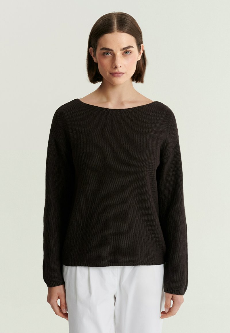Femme aux cheveux courts et bruns portant un pull en maille noir et un pantalon blanc, debout devant un fond clair uni.