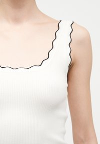 Λευκό ριμπ τοπ με κυματιστές άκρες που έχουν μαύρο περίγραμμα, φορεμένο από ένα άτομο, εμφανίζοντας τους ώμους και το άνω μέρος του στήθους.