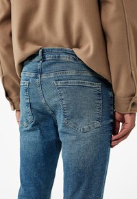 Vaqueros de mezclilla azul con un corte de altura media, que cuentan con dos bolsillos traseros y una textura desgastada. Una blusa beige cubre parcialmente la cinturilla.