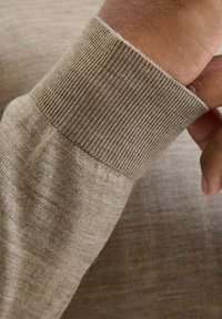 Ljusbrun stickad tröja i detalj som visar ribbad mudd och slät textur, hand delvis synlig som greppar tyget.