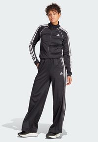 Black athletic tracksuit made of smooth fabric, featuring white Adidas stripes along the sides and a zip-up jacket with a high collar.
