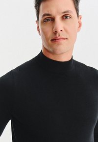Czarny sweter z golfem, z prążkowanym dekoltem, dopasowaną konstrukcją, gładką teksturą oraz bliskim fasonem, noszony przez modelkę.