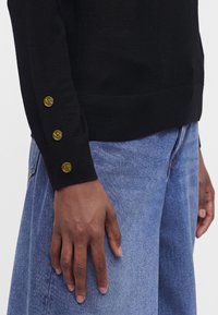 Personne portant un haut noir à manches longues avec trois boutons dorés sur le poignet et un jean bleu, main détendue le long du corps.