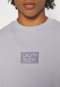 Ljus lavendelfärgad sweatshirt med korta ärmar, ribbad rundhalsning och en "Calvin Klein"-logotyp i en rektangulär, lila-vit design på framsidan.