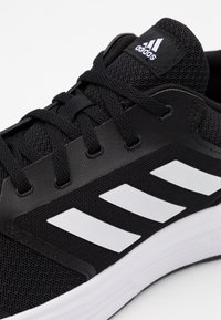 Svart träningsträskor med mesh-överdel, platta snören och tre vita ränder på sidan. Har en texturerad design och dämpad sula.