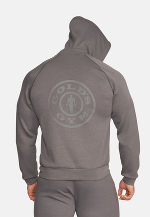 Mann in grauem Hoodie und Hose, von hinten gesehen, mit dem Gold's Gym-Logo zentral auf dem Rücken des Hoodies.