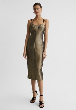 Frau trägt ein figurbetontes, midilanges Kleid in metallischem Gold mit schwarzen dünnen Trägern und schwarzen Riemchen-High-Heel-Sandalen, steht vor einem schlichten Hintergrund.