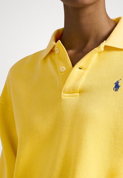 Osoba w żółtej koszulce polo z zapiętymi na dwa guziki i małym niebieskim haftowanym logo na lewej piersi.