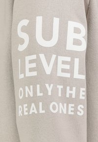 Beige stof met witte tekst die "SUB LEVEL ONLY THE REAL ONES" in vetgedrukte, hoofdletters weergeeft.