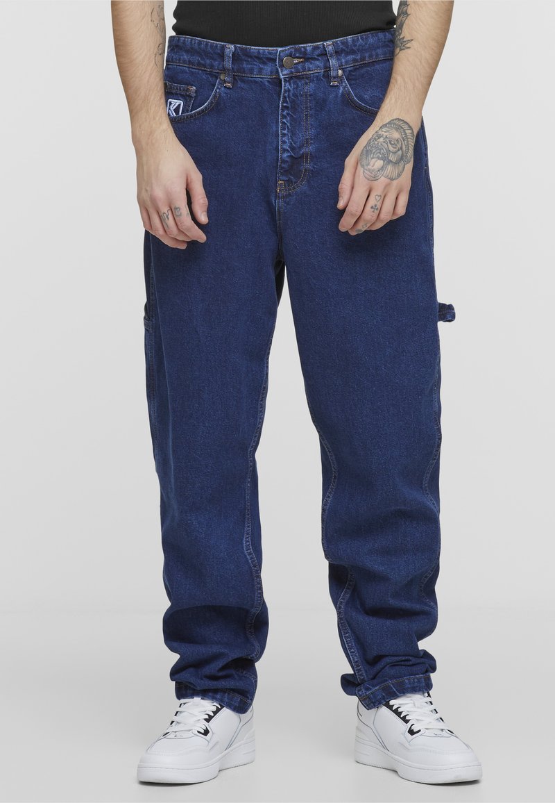 Mann trägt lockere dunkelblaue Jeans aus Denim und weiße Turnschuhe, Hände sichtbar mit Tätowierungen auf Fingern und Handgelenk.