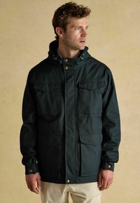 Jachetă verde închis cu glugă, având două buzunare pe piept și închideri cu capse. Materialul pare neted, cu un aspect mat și cu accente din metal auriu.