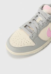Gros plan d'une basket grise et crème avec un logo rose, une boîte à orteils perforée et des lacets gris sur un fond clair.