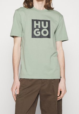 Personne portant un t-shirt vert clair à manches courtes avec un grand logo carré noir portant la mention "HUGO" en lettres majuscules, associé à un pantalon marron.