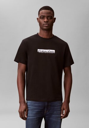 Ung mann iført en svart Calvin Klein T-skjorte og blå jeans, stående mot en ensfarget grå bakgrunn, vendt mot fronten.