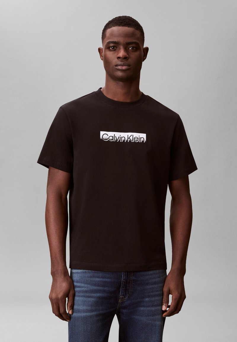 Junger Mann trägt ein schwarzes Calvin Klein T-Shirt und eine blaue Jeans, steht vor einem schlichten grauen Hintergrund und schaut geradeaus.