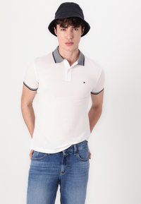 Hvit polo skjorte med grå krage og erme aksenter, med en liten logo. Kombinert med blå denim jeans og en svart bøttehatt.